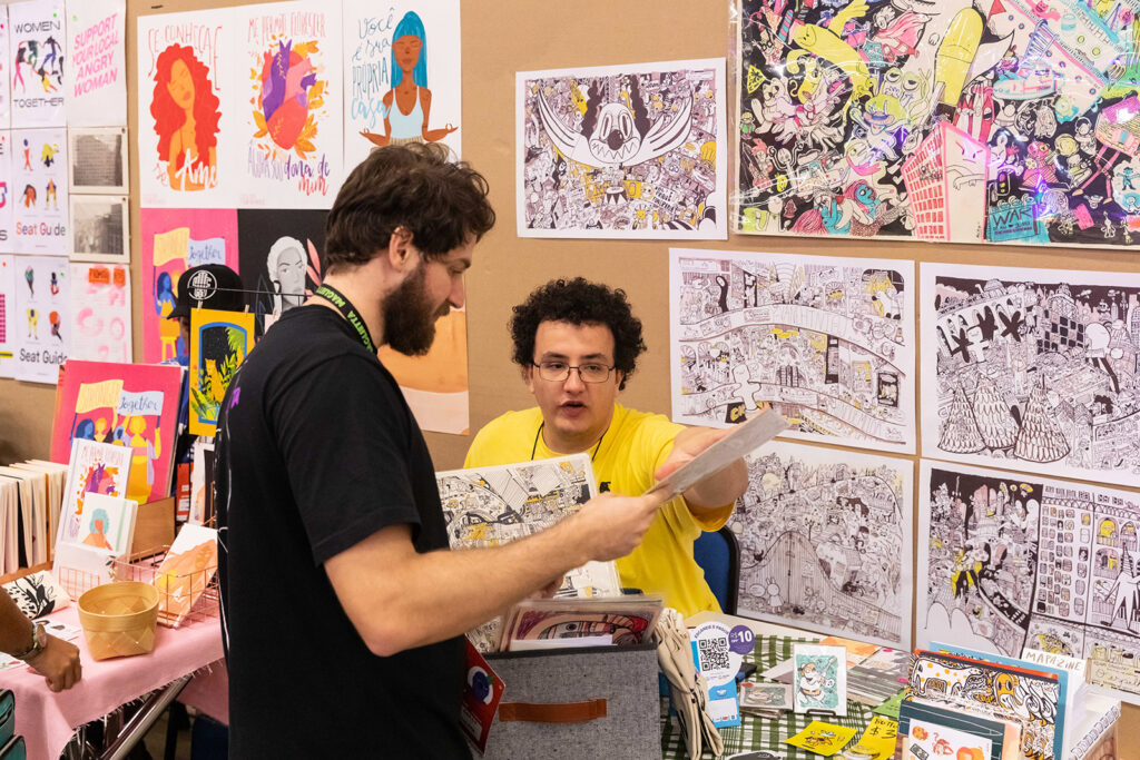 Feira com estandes de arte e publicações, com participantes interagindo e selecionando obras durante o Pixel Show.