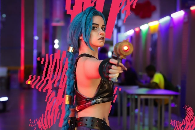 Pessoa com cabelo azul longo caracterizada como personagem de videogame, usando roupa preta e apontando uma arma cenográfica em direção à câmera. O ambiente ao fundo tem iluminação colorida típica de evento ou convenção geek.