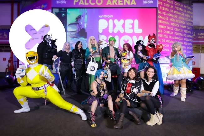 Grupo de pessoas vestidas como personagens de games, anime e cultura pop posando para foto em frente ao palco do evento Pixel Show. Os figurinos incluem armaduras, vestidos temáticos e um personagem com traje amarelo inspirado em super-herói.