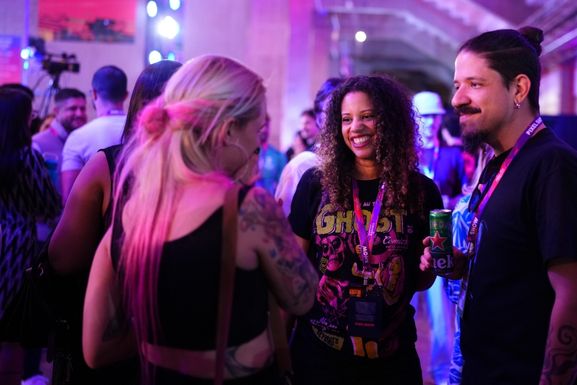 Três pessoas conversam e sorriem em um ambiente de evento com iluminação colorida roxa e azul. Uma mulher com cabelo cacheado usa camiseta preta e crachá do evento, enquanto conversa com uma mulher loira de cabelo preso que tem tatuagens no braço. Ao lado, um homem com cabelo preso em coque e barba segura uma lata de bebida enquanto participa da conversa. Ao fundo, outras pessoas circulam pelo espaço do evento.