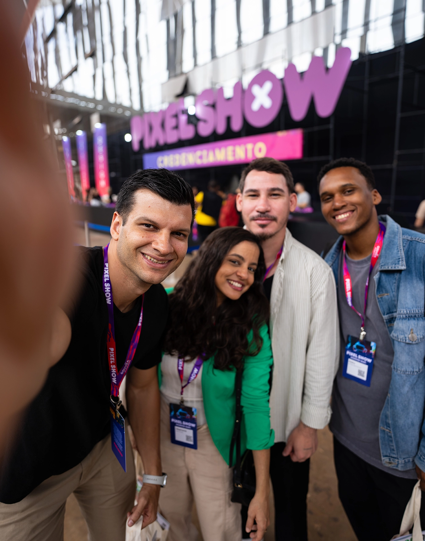 Quatro participantes do Pixel Show, de diferentes perfis, posam sorrindo para uma selfie dentro do evento, usando credenciais no pescoço, com o logo Pixel Show visível ao fundo, representando a comunidade criativa de Brasília.